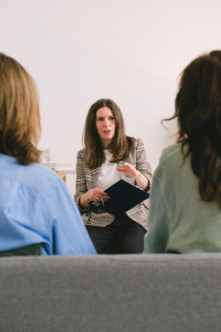 Pensive woman entrepreneur with brown hair in casual clothes sitting with clipboard and talking with unrecognizable clients in light room