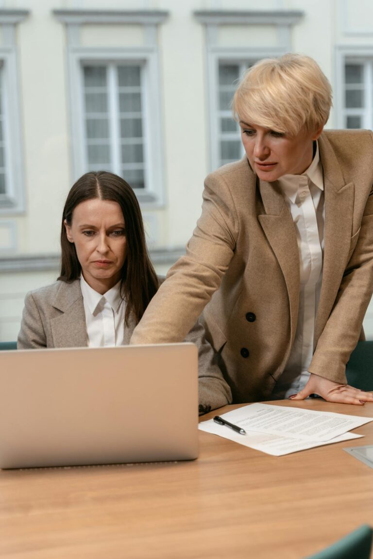 Two businesswomen collaborating at a desk in a modern office, focused on a laptop.